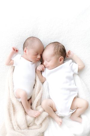 Two newborn twins sleeping peacefully side by side in white onesies on a soft cream blanket.