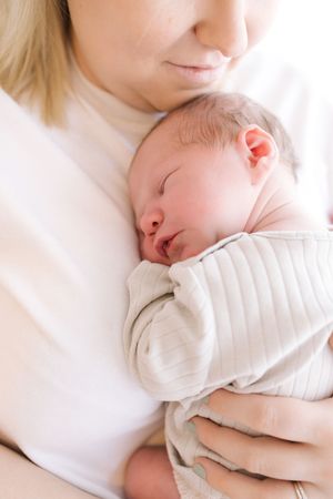 A mother tenderly holds her sleeping newborn baby wrapped in a soft white swaddle blanket close to her chest.
