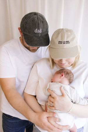 New parents wearing dad and mum baseball caps tenderly hold their sleeping newborn baby together.