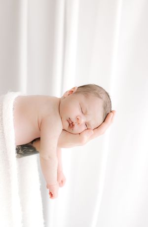 Sleeping newborn baby cradled in a tattooed hand on a soft white blanket.