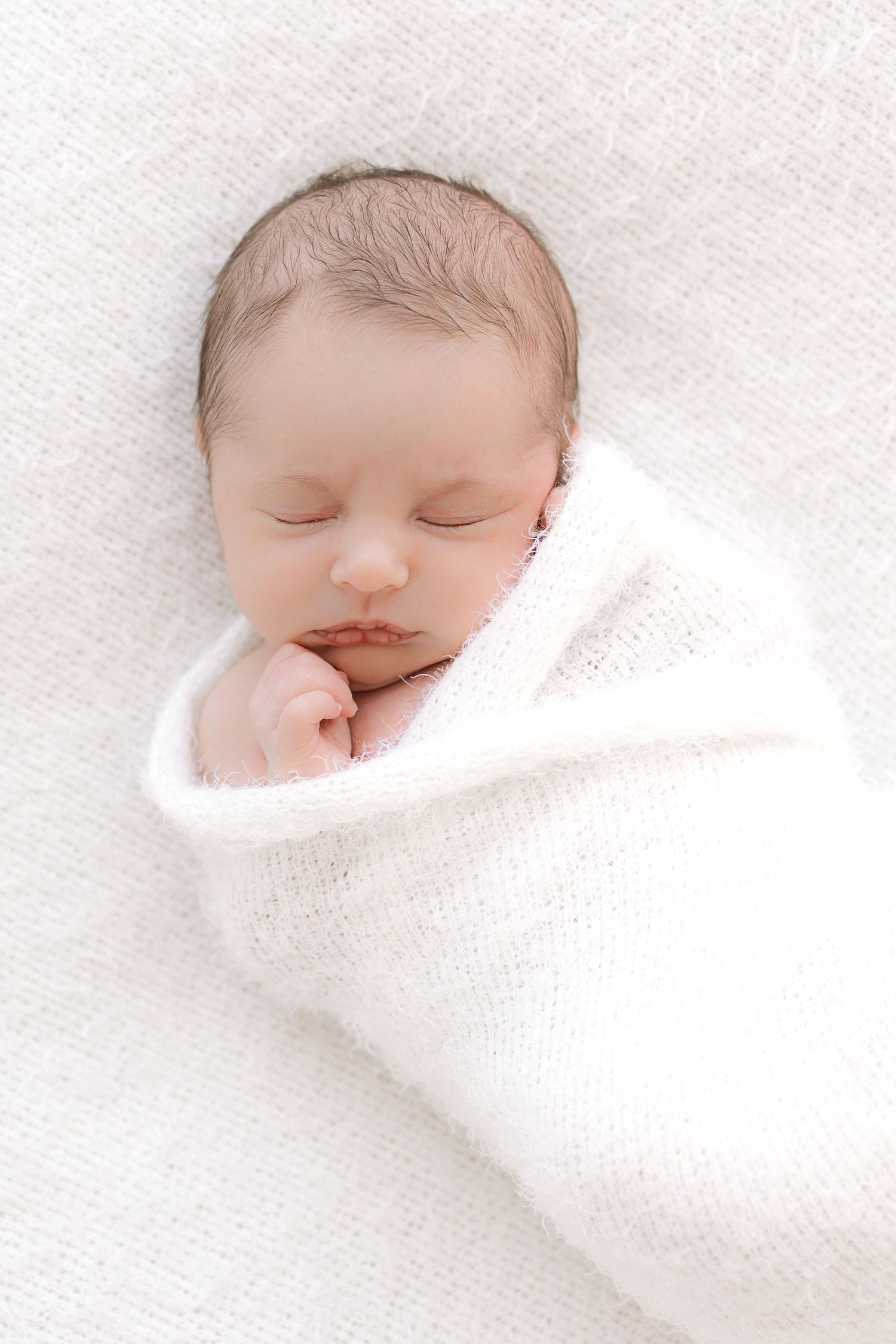 Peaceful newborn baby swaddled in a soft white knit blanket, sleeping with tiny hand near face.