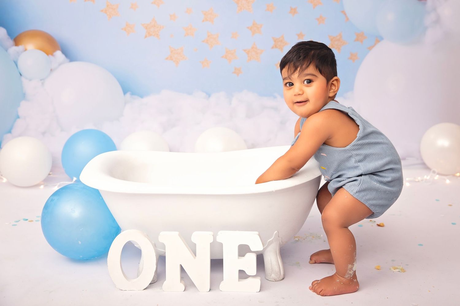 Cute toddler boy in blue romper stands by white bathtub during ONE first birthday photo session.