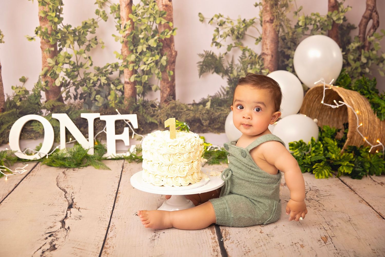 Baby boy in green romper sits with white cake during woodland themed first birthday photo session.