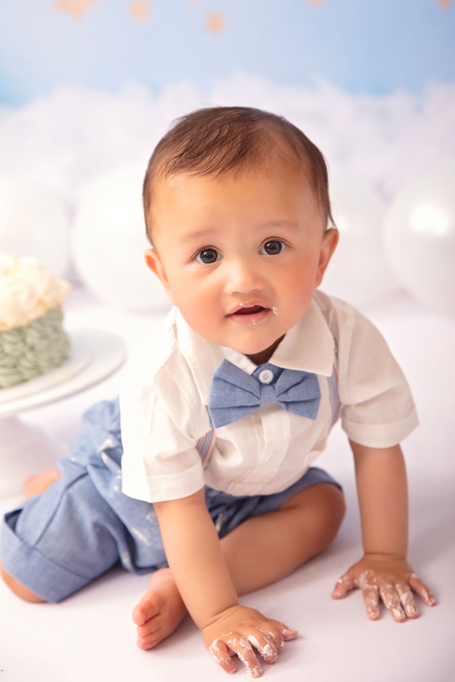 Adorable baby boy in white shirt and blue bow tie sits for his first birthday cake smash photo session.