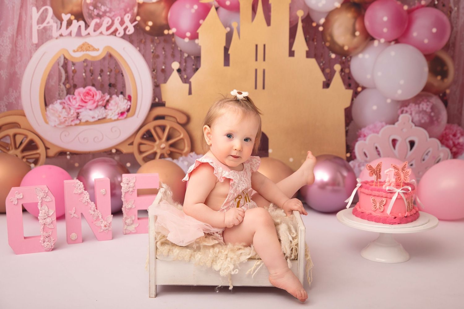Baby girl in pink romper sits on mini bed at princess castle themed first birthday photo session.