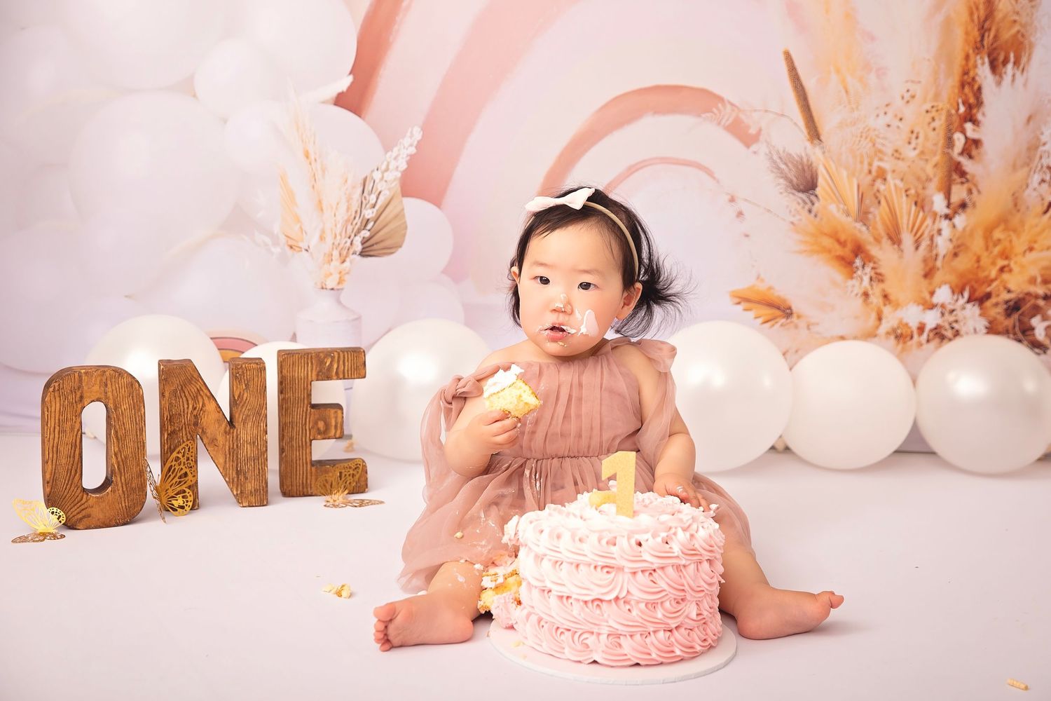 Baby girl in pink dress smashes her first birthday cake surrounded by boho rainbow balloon backdrop.