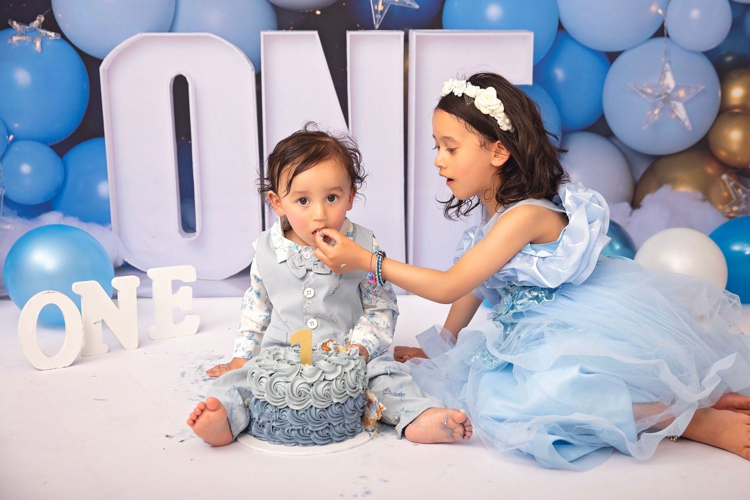 Older sister in blue dress feeds baby brother cake during his first birthday ONE cake smash session.