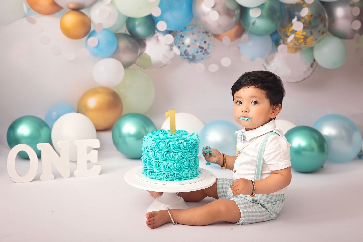 Baby boy in plaid outfit sits with teal rosette cake during colorful balloon first birthday session.