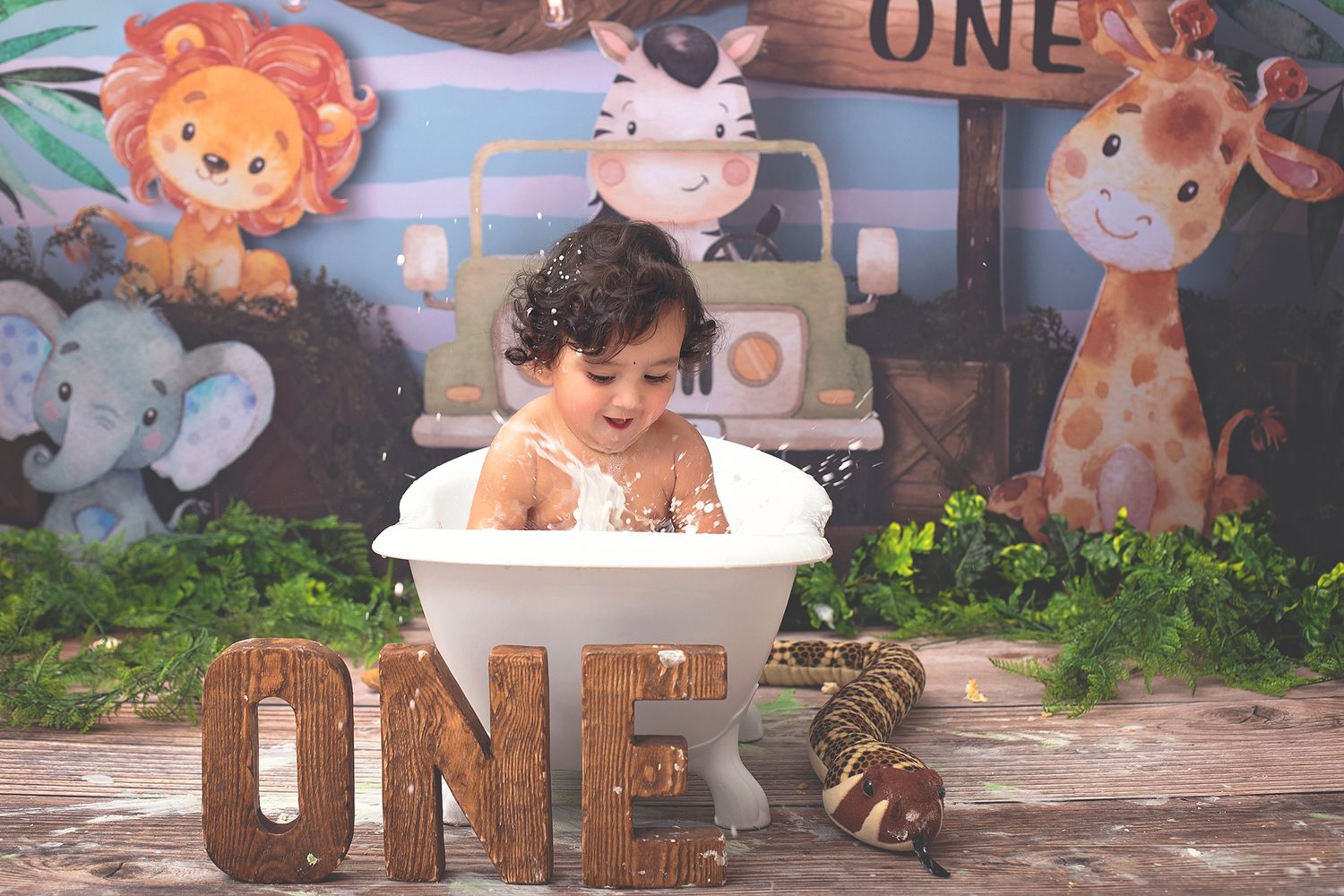 Smiling one-year-old splashing in a white mini bathtub with colorful safari animal birthday party backdrop.