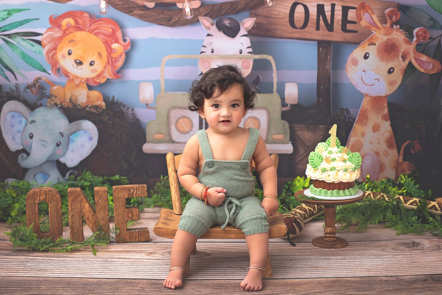 Adorable one-year-old boy in green overalls sitting on a bench with safari backdrop and birthday cake.