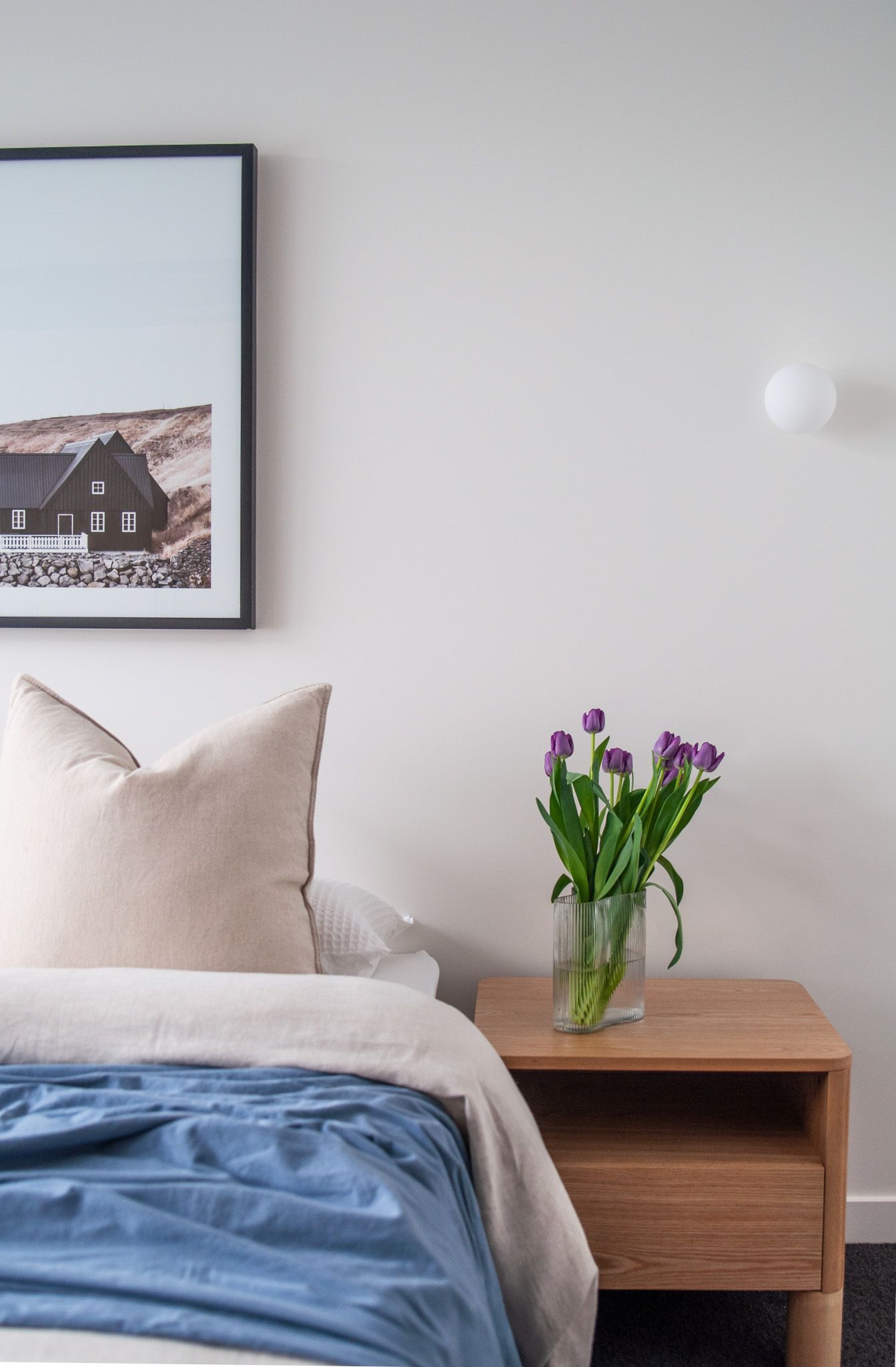 Cozy bedroom corner with framed house print, purple tulips, and blue bedding.