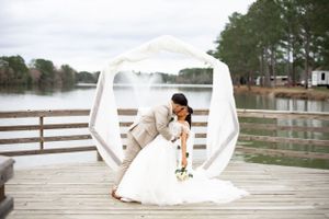Artistic wedding dip pose under white arch on lakeside wooden dock.