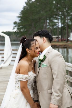 Intimate moment between bride in off-shoulder dress and groom in beige suit on lakeside pier.