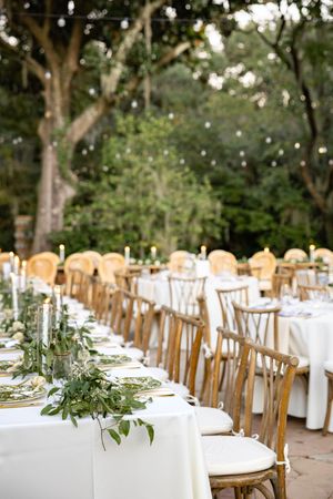Elegant outdoor wedding reception setup with wooden chairs and white tablecloths under a canopy of trees.
