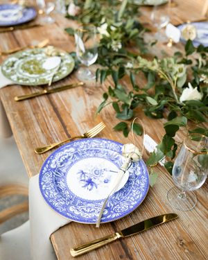 Blue and white patterned china plates with gold utensils are set on a rustic wooden table with scattered greenery.