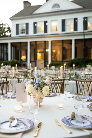 Elegant outdoor wedding reception setup with blue and white china, candlelight, and a grand white colonial house in background.