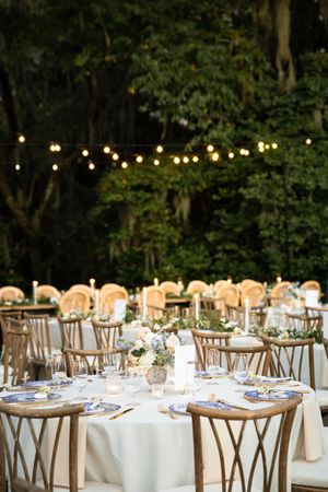 Elegant outdoor wedding reception tables with white linens, wooden chairs, and string lights hanging in trees above.