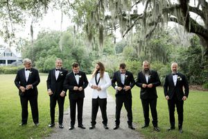 Groomsmen in black tuxedos stand in line under Spanish moss draped oak trees at an outdoor wedding venue.