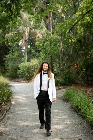 A person in formal black and white attire walks along a scenic gravel path surrounded by lush green foliage.