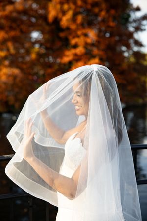 A bride in a white wedding dress lifts her delicate veil against a backdrop of autumn trees with orange foliage.