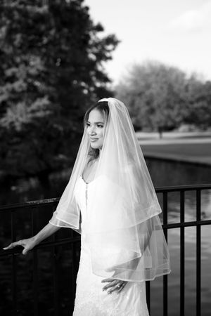 A bride in a flowing white wedding dress and veil poses elegantly against a railing in black and white photography.
