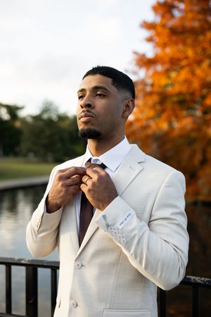 Person in white suit adjusts their collar against autumn foliage backdrop near waterfront railing at sunset.