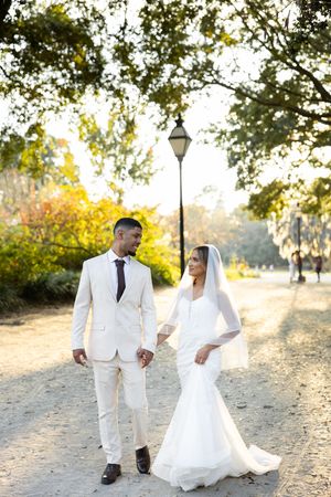 A couple in elegant white wedding attire poses together on a sunlit path surrounded by autumn trees.