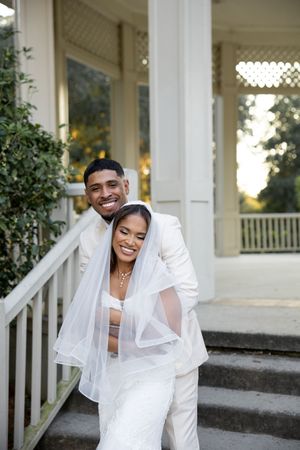Joyful couple in white wedding attire share playful poses on a white porch during their wedding photoshoot.