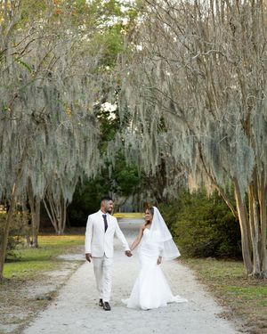 A couple in white attire walks through a scenic path lined with Spanish moss-draped trees at an outdoor venue.