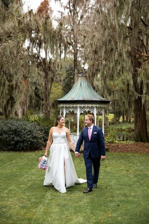 A couple walks together on a lush green lawn with Spanish moss-draped trees and a gazebo in the background.