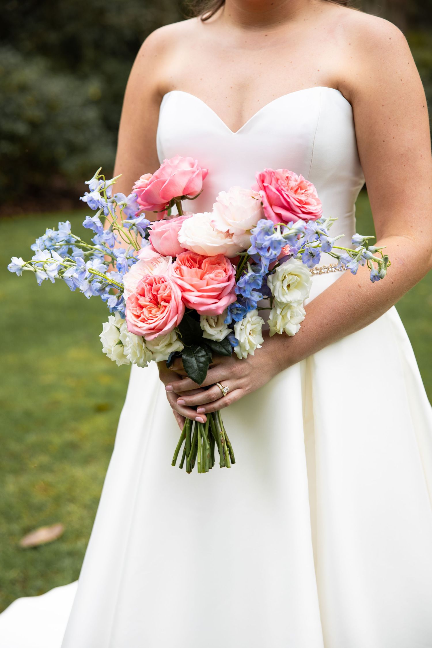 Beautiful wedding bouquet featuring coral pink roses and delicate blue flowers against a white dress.
