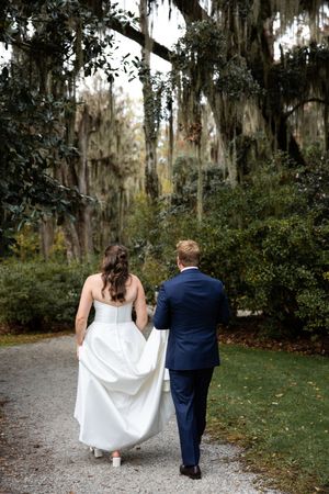 A couple walks down a Spanish moss-draped path in formal wedding attire at a Southern garden venue.