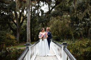 Wedding couple poses on white wooden bridge surrounded by lush greenery and Spanish moss in romantic garden setting.