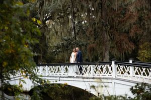 A couple walks across a white ornate bridge in a lush garden setting during a sunny day.