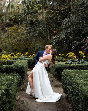 Romantic garden wedding scene with couple embracing among green hedges and yellow flowers.