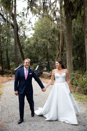 Newlyweds share a joyful moment walking hand-in-hand along a tree-lined gravel path.