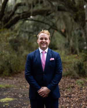 A groom in a navy suit with pink tie poses for portraits in a wooded outdoor setting.
