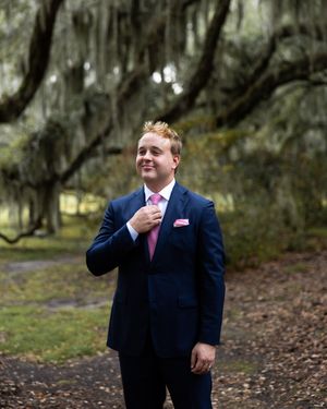 A groom in navy suit with pink tie adjusts his collar under moss-draped trees.