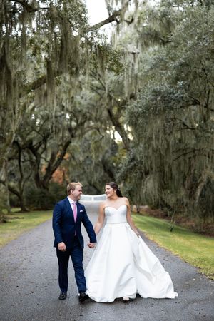 Wedding couple strolls along a gravel path beneath Spanish moss-covered oak trees.