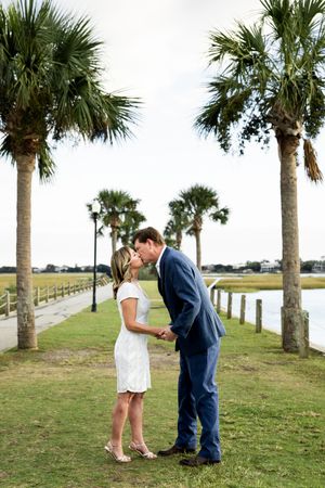 A couple shares a romantic moment on a palm tree-lined path by the water during a sunset proposal.