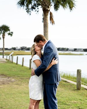 A couple shares a romantic moment by the waterfront with palm trees and a wooden fence in the background.