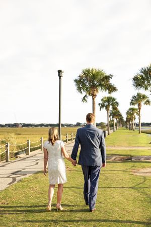 A couple walks down a palm tree-lined pathway at sunset on a grassy coastal area.