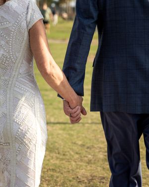 A couple in formal attire holds hands while standing on grass during an outdoor wedding ceremony.