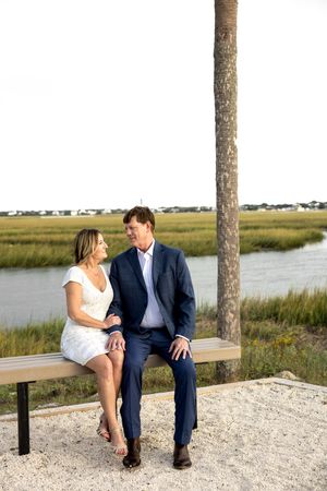 A couple sits together on a bench overlooking a scenic coastal marsh landscape on a sunny day.