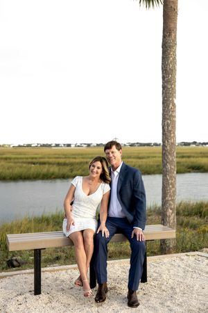 A couple sits together on a bench overlooking a scenic coastal marsh landscape during sunset.