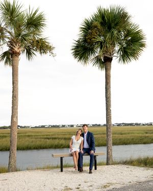 Romantic coastal scene with palm trees lining a sandy beach path beside tranquil marshland at sunset.