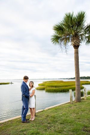 A couple shares an intimate moment by the water's edge with palm trees and marsh grass along the coastline.