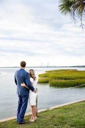 Two people stand together on a grassy waterfront overlooking a coastal marsh landscape with water views.