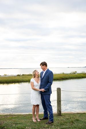A couple stands together by a waterfront with marsh grass and cloudy skies in the background.