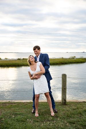 A couple shares a romantic moment by the water's edge at sunset with marsh grass and cloudy skies in the background.